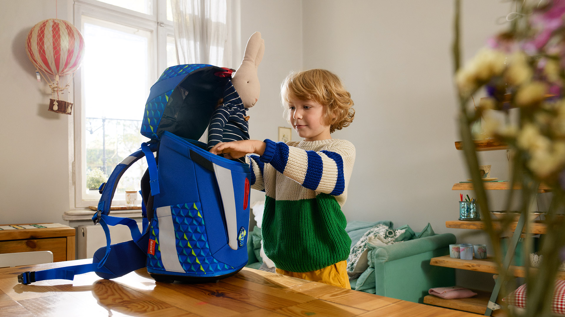 Junge packt sein Stofftier in einen blauen Schulranzen ein.